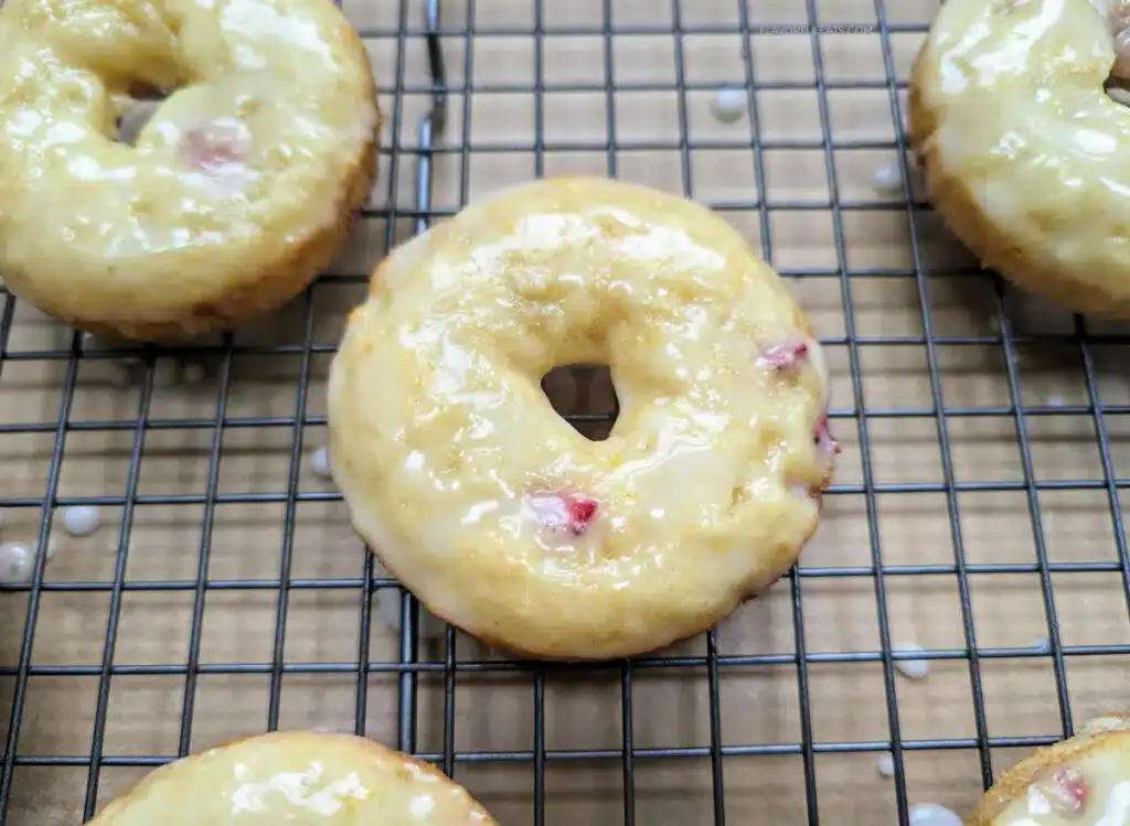 A glazed baked donut with small red fruit pieces sits on a wire cooling rack.