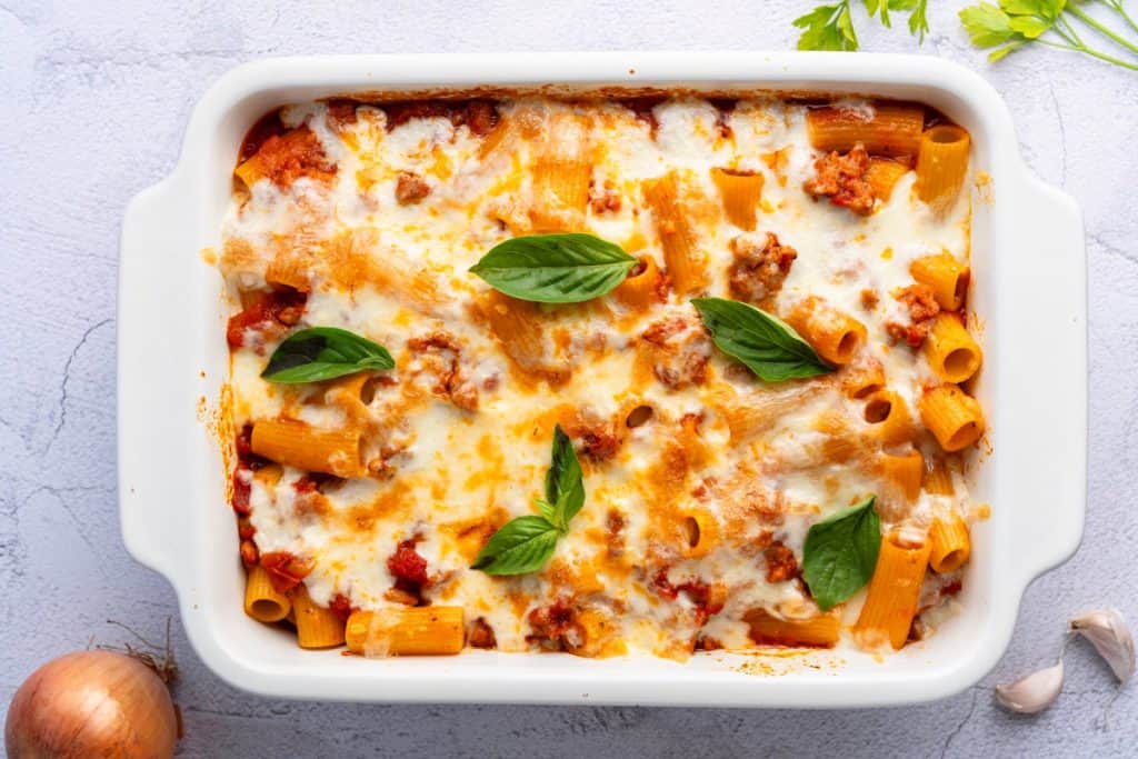 A baked pasta dish with rigatoni, melted cheese, tomato sauce, ground meat, and basil leaves in a white casserole dish.