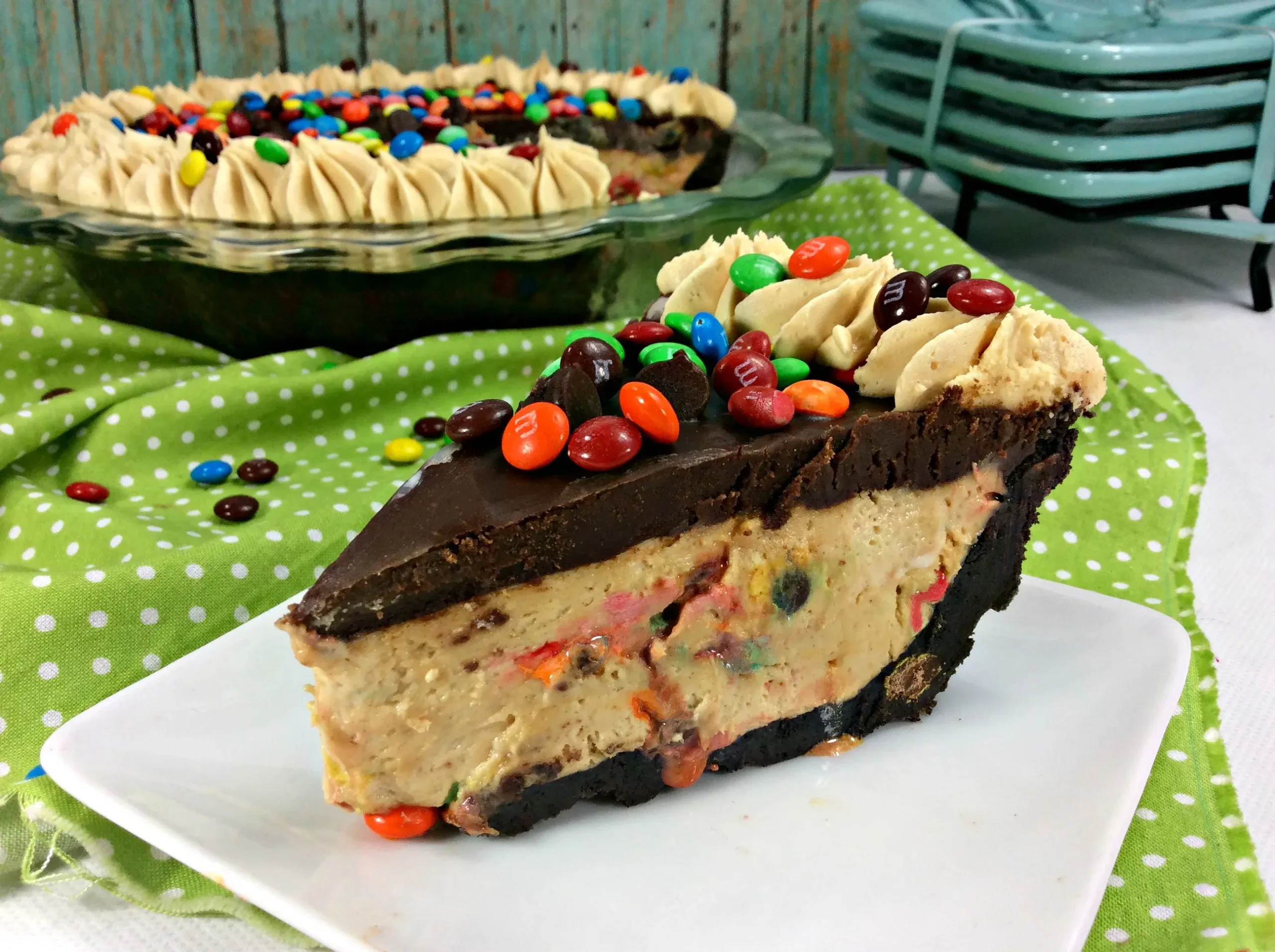 A slice of chocolate peanut butter pie with an Oreo crust, topped with frosting and colorful candy-coated chocolates, sits on a white plate in front of the whole pie.