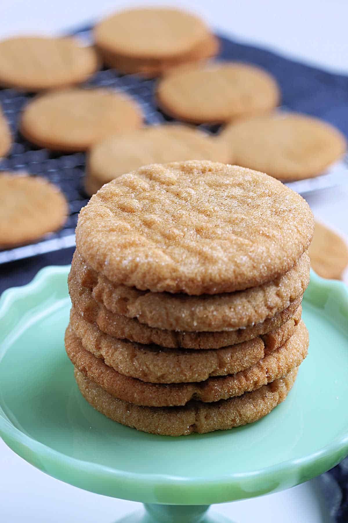A stack of peanut butter cookies sits on a green cake stand, with more cookies cooling on a wire rack in the background.