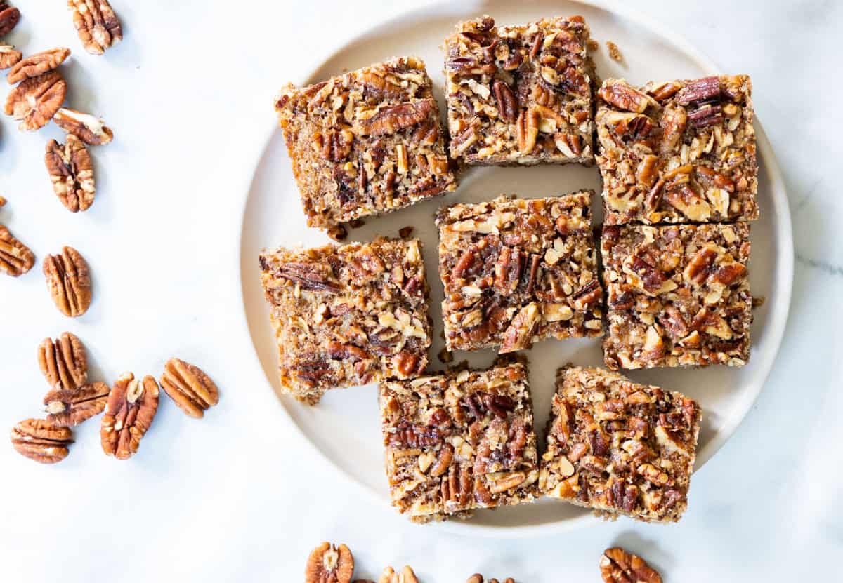 A plate with seven pecan bars cut into squares, surrounded by scattered pecan halves on a white surface.
