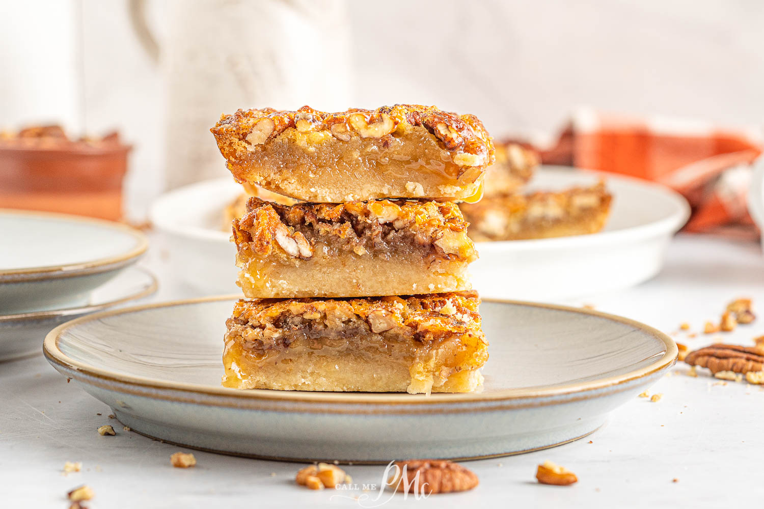 Three pecan pie bars are stacked on a plate, showing a layered filling with chopped pecans on top. A few pecan pieces are scattered around the plate.