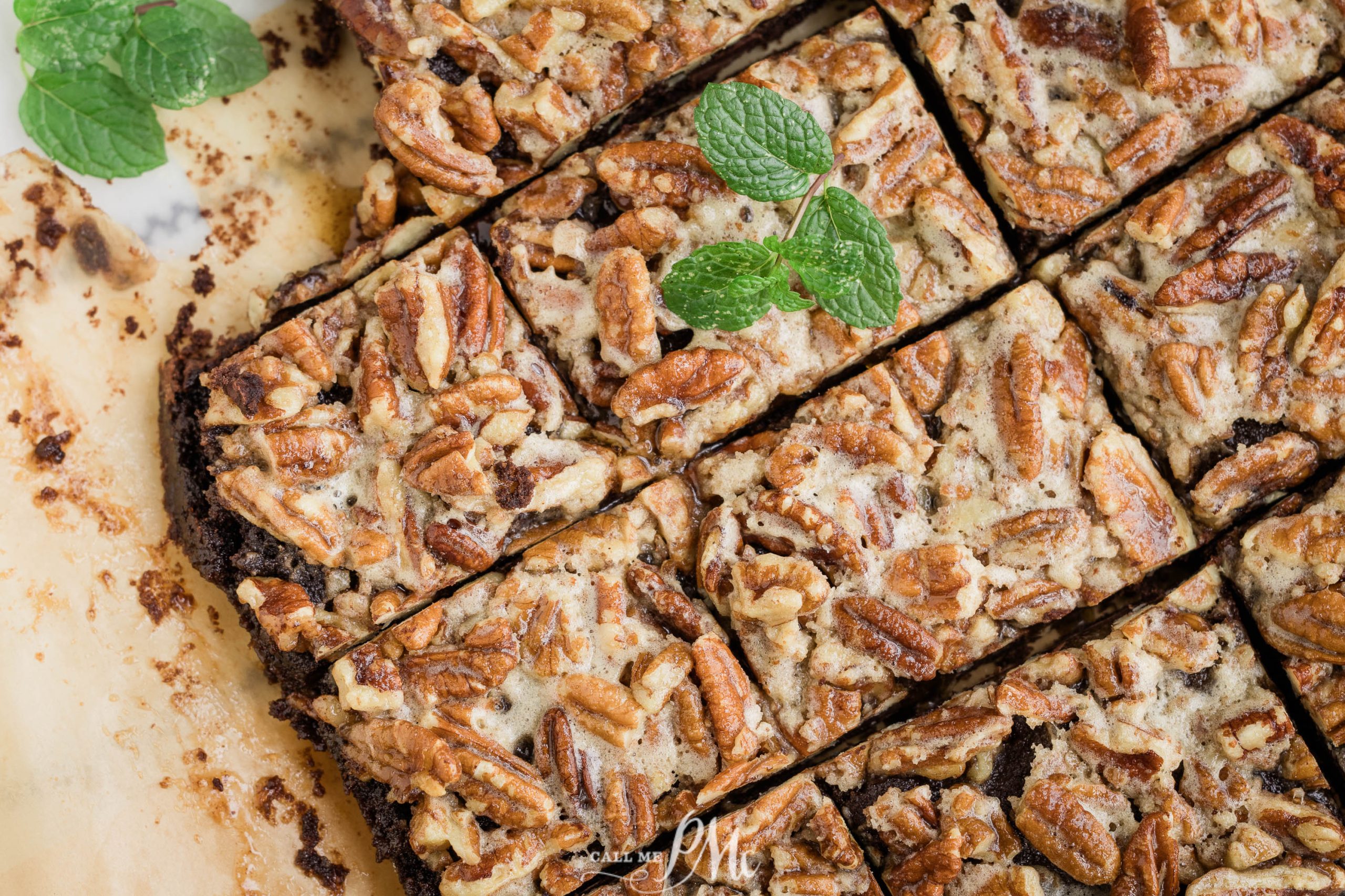 Close-up of sliced pecan-topped dessert bars garnished with fresh mint leaves on a parchment-lined surface.