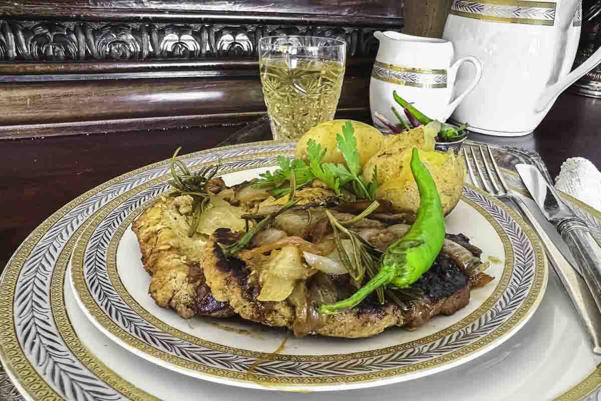 A plated meal with grilled meat topped with onions and herbs, roasted potatoes, a green chili pepper, and a glass of light-colored beverage in the background.