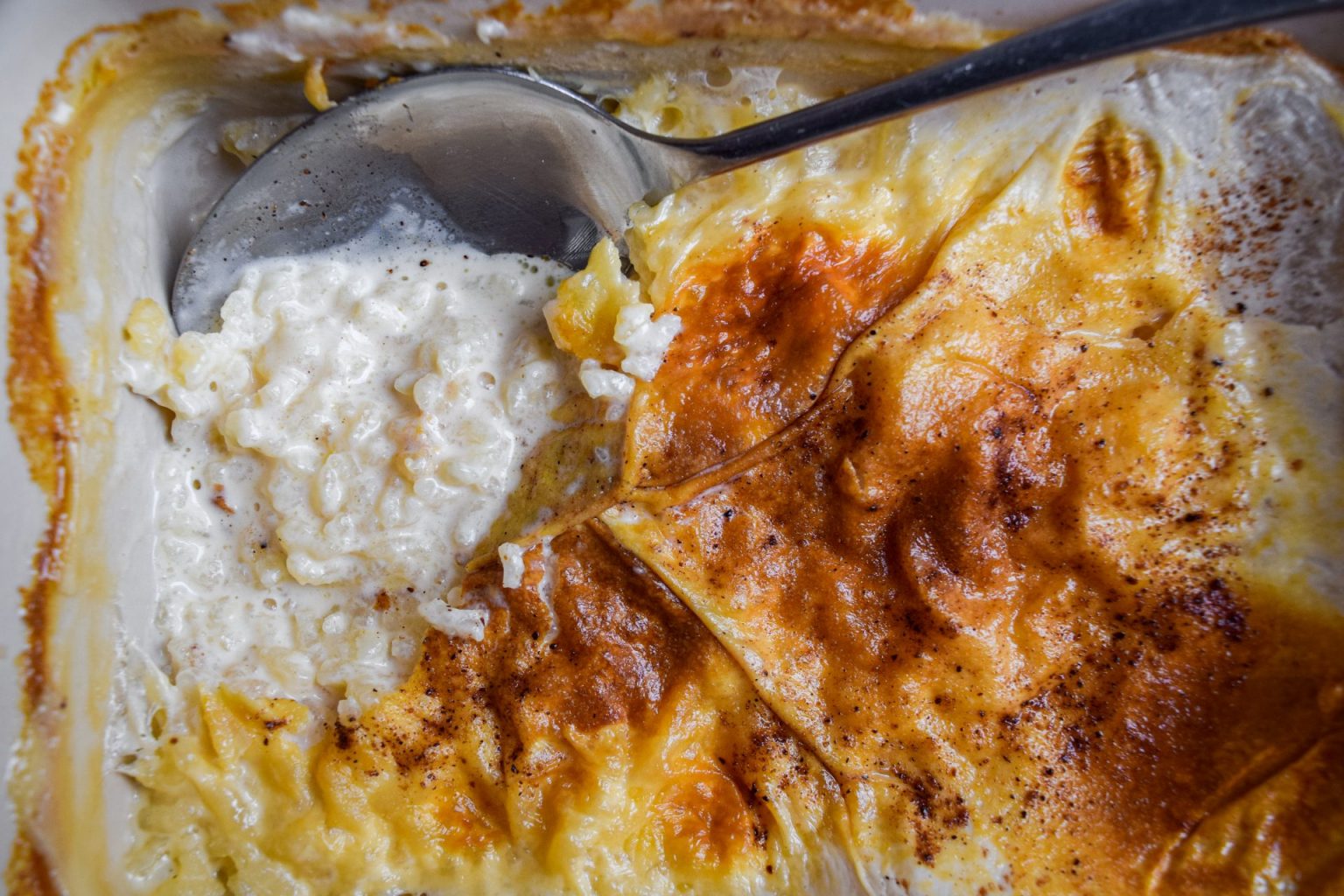 A close-up of a dish of baked rice pudding with a browned, custard-like top and a serving spoon partially submerged in the creamy mixture.