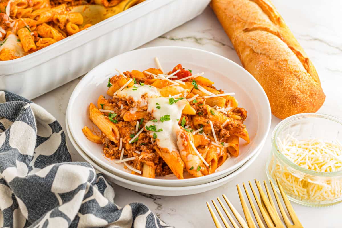 A plate of baked penne pasta with meat sauce and melted cheese, served with a baguette, grated cheese, and gold forks on a marble surface.