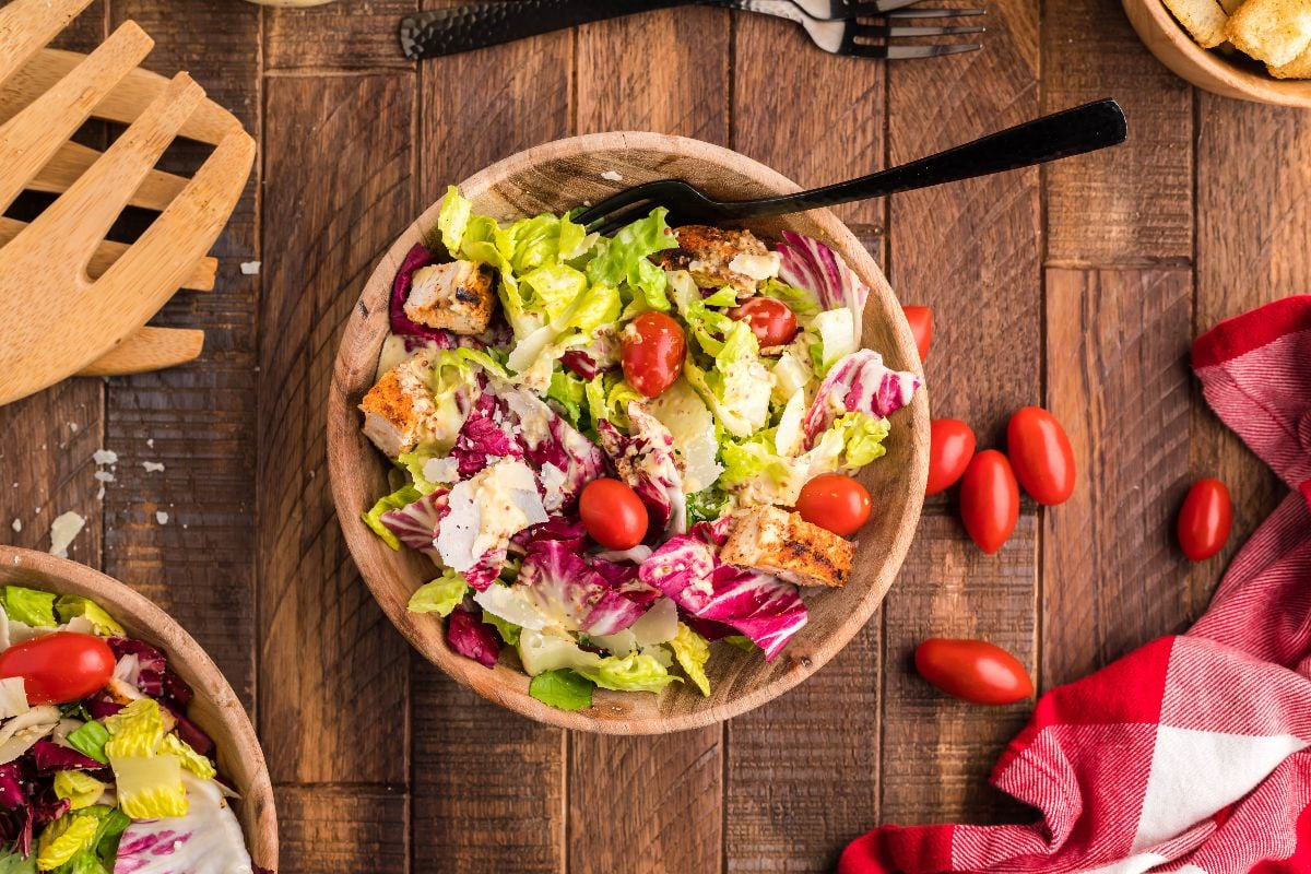 A wooden bowl of salad with lettuce, cherry tomatoes, croutons, and cheese on a wooden table, next to utensils and a red checkered napkin.