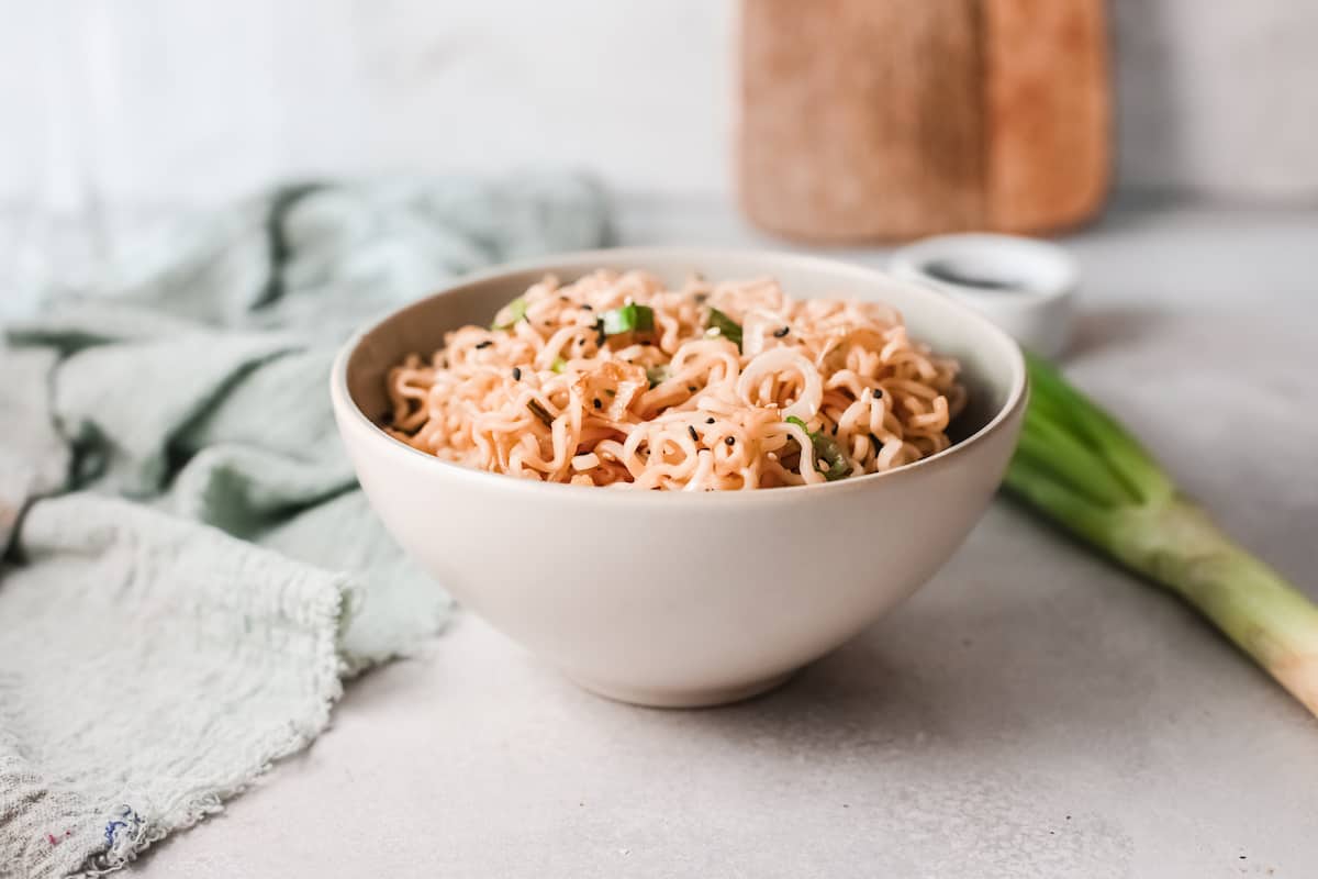A white bowl filled with cooked instant noodles garnished with green onions sits on a light countertop next to a green onion and a cloth.
