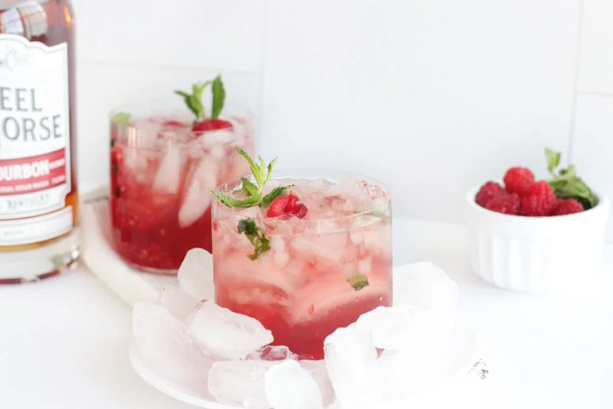 Glass of pink cocktail with ice, mint, and raspberries on a white plate with ice cubes. A bottle of bourbon and a bowl of raspberries are in the background.