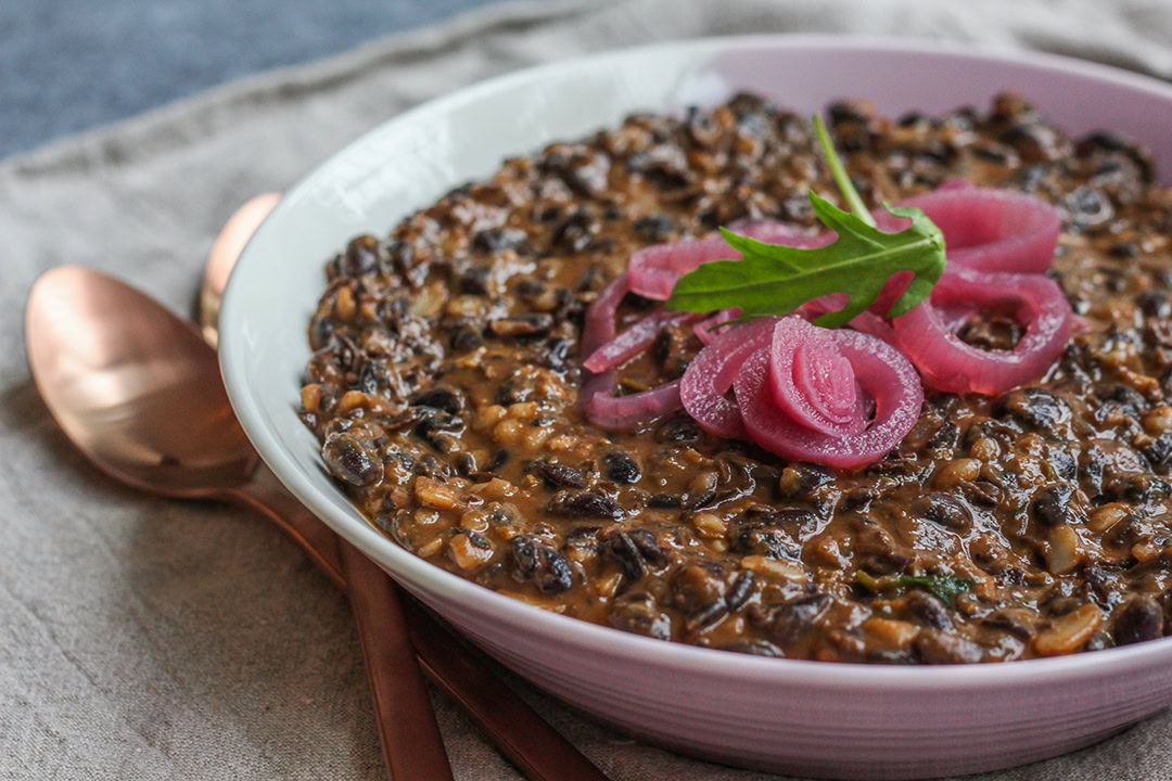 A bowl of creamy mushroom risotto topped with pickled red onions and a green leaf, placed next to copper-colored utensils on a beige cloth.