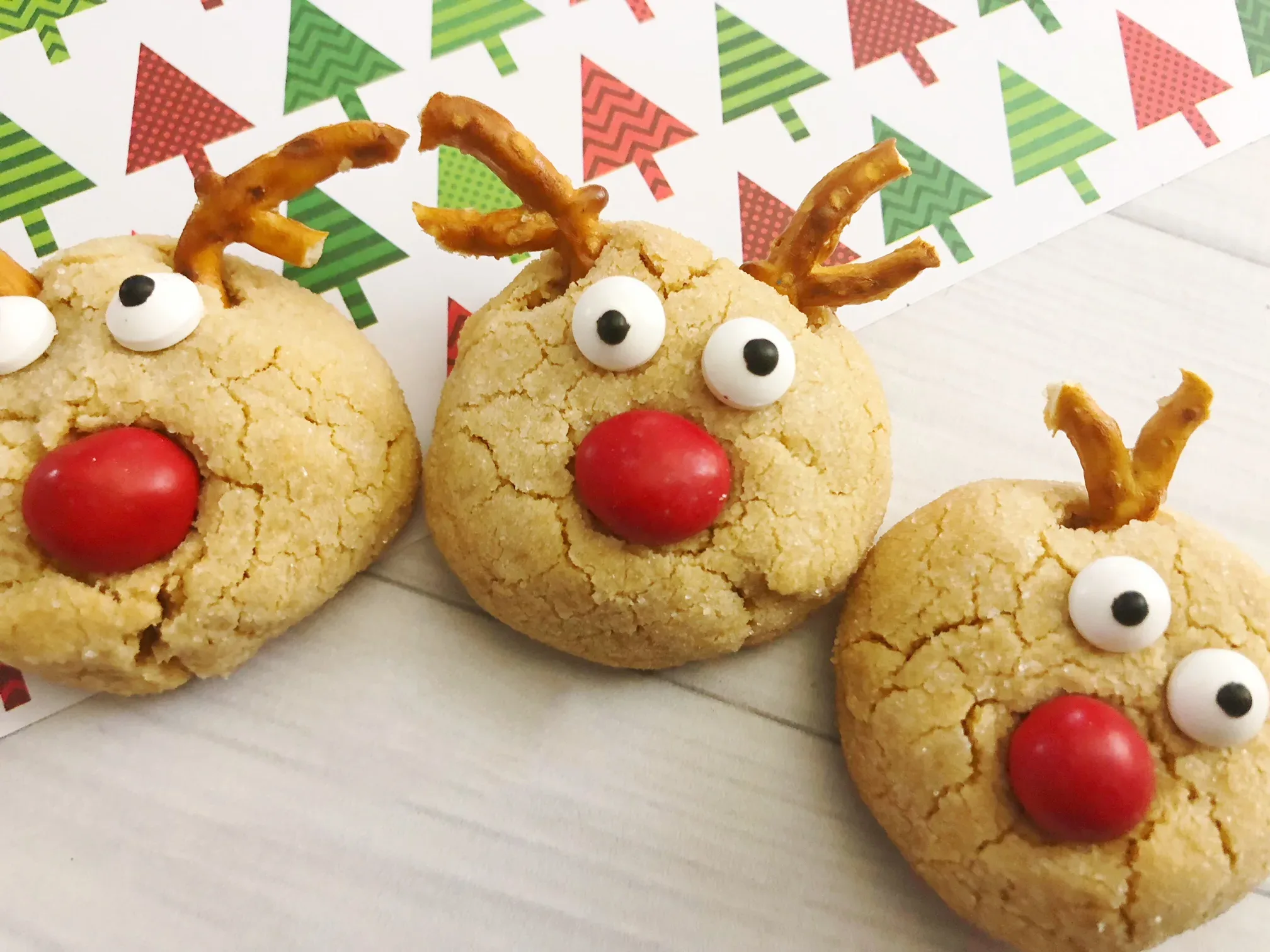 Three cookies decorated as reindeer with candy eyes, red candy noses, and pretzel antlers on a white surface with a Christmas tree patterned paper.