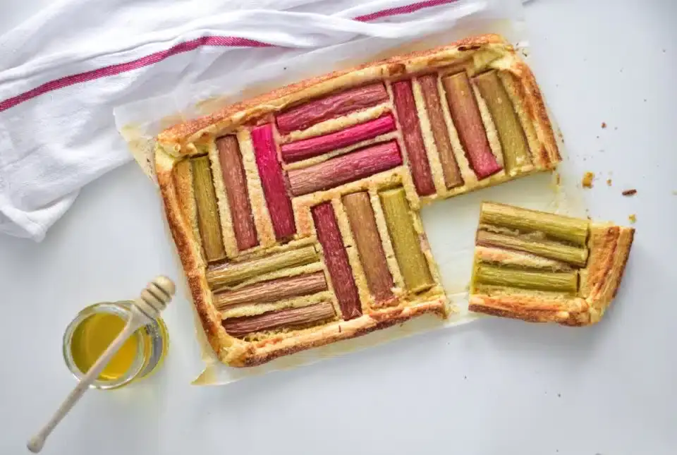 Rectangular rhubarb tart with geometric pattern on a white surface, one slice cut out, and a jar of honey with a dipper nearby.