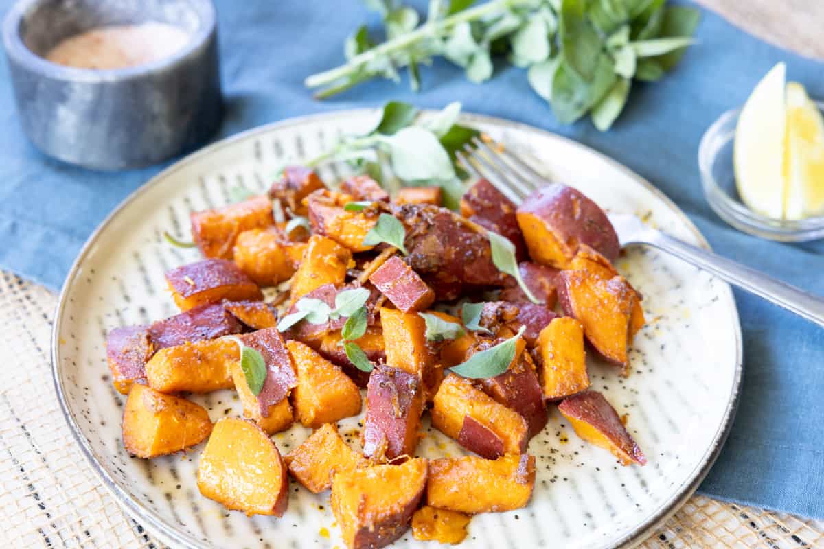 A plate of roasted sweet potato cubes garnished with fresh herbs, with a fork, a lemon wedge, and a small bowl of seasoning on the side.