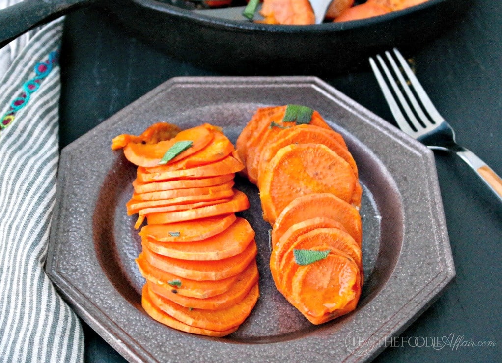 Sliced, cooked sweet potatoes are arranged neatly on a brown plate, garnished with small green herb leaves. A fork and striped napkin are nearby.