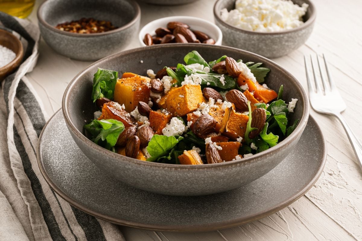 A bowl of salad with roasted pumpkin, leafy greens, almonds, and crumbled cheese, with small bowls of toppings and a fork nearby.
