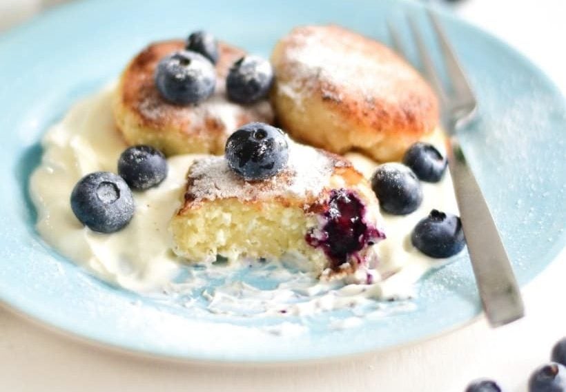 Three cottage cheese pancakes with a dollop of cream and fresh blueberries on a blue plate, one pancake is partially eaten, fork on the side.