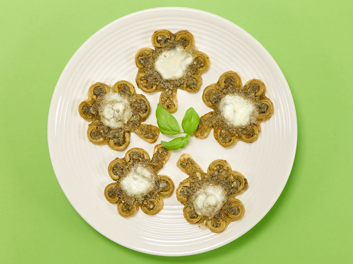 Five clover-shaped savory waffles with melted cheese on top are arranged on a white plate, garnished with fresh basil leaves, against a green background.