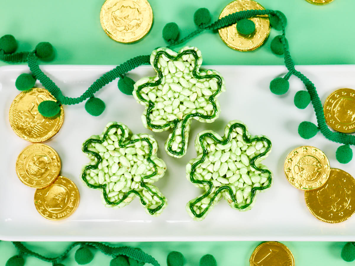 Three shamrock-shaped treats made of green and white candies on a white tray, surrounded by gold chocolate coins and green pom-pom garland on a light green background.