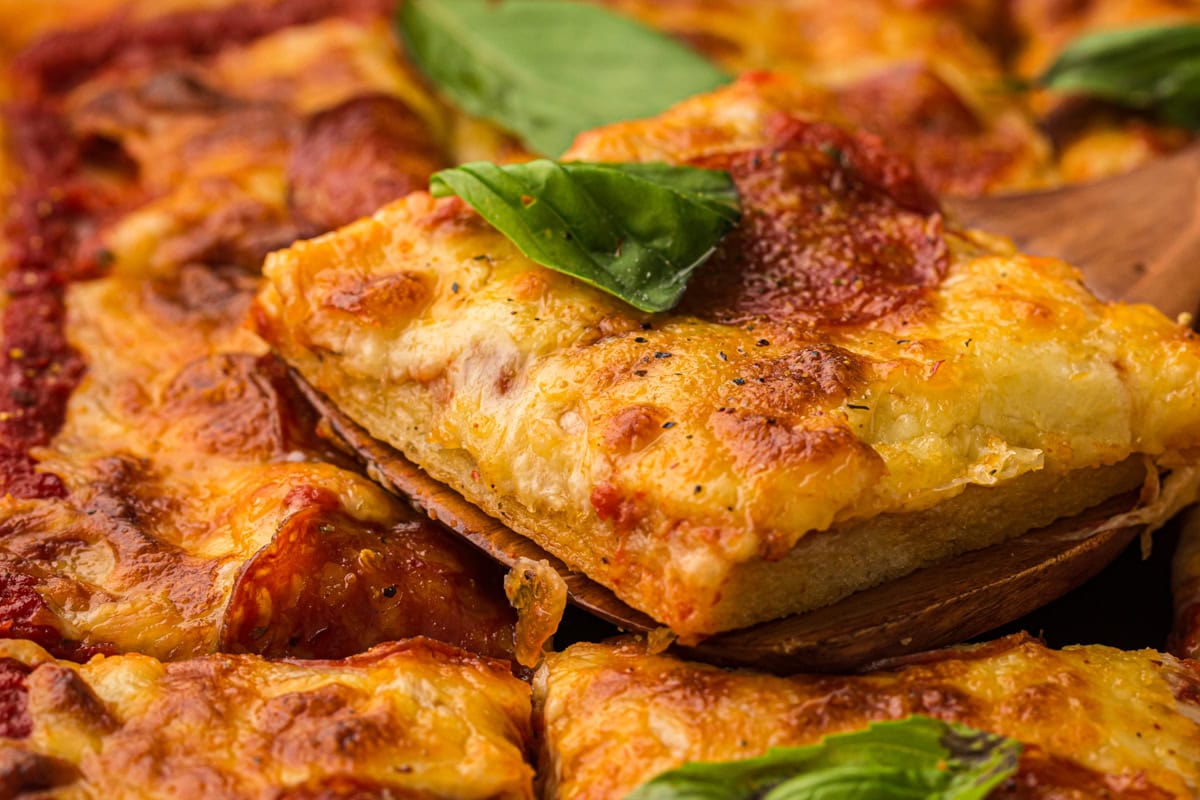 A close-up of a rectangular slice of pepperoni pizza being lifted from a tray, topped with melted cheese and fresh basil leaves.