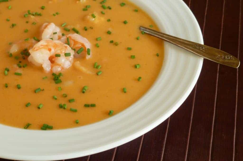 A bowl of creamy orange-colored bisque topped with shrimp and chopped chives, with a spoon resting on the rim of the white bowl.