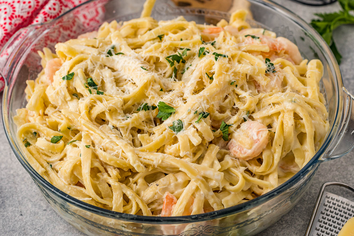 A glass dish filled with fettuccine pasta in a creamy white sauce, topped with grated cheese, chopped parsley, and pieces of shrimp.