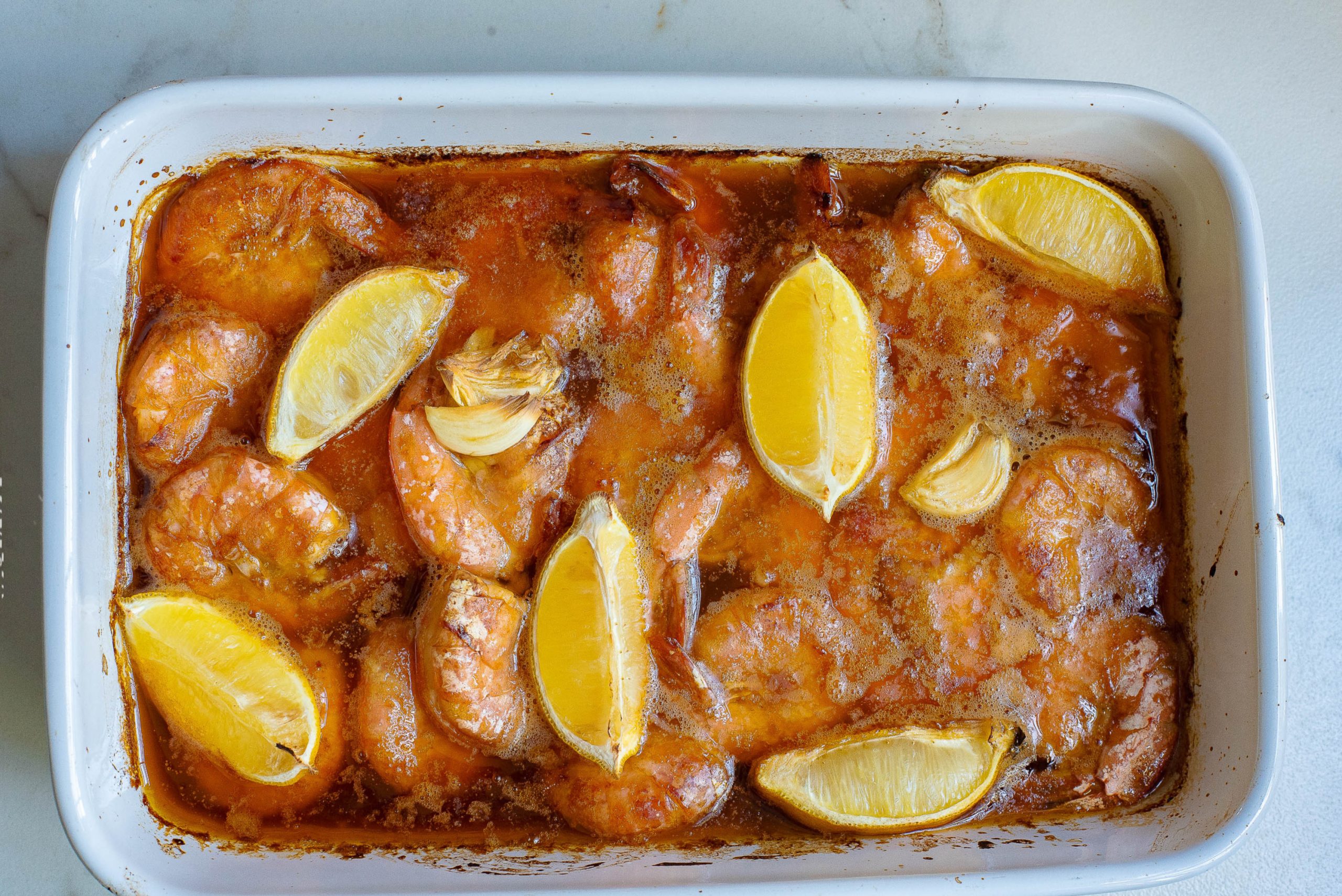 Baked shrimp in a tomato-based sauce topped with lemon wedges and garlic cloves in a white rectangular dish.