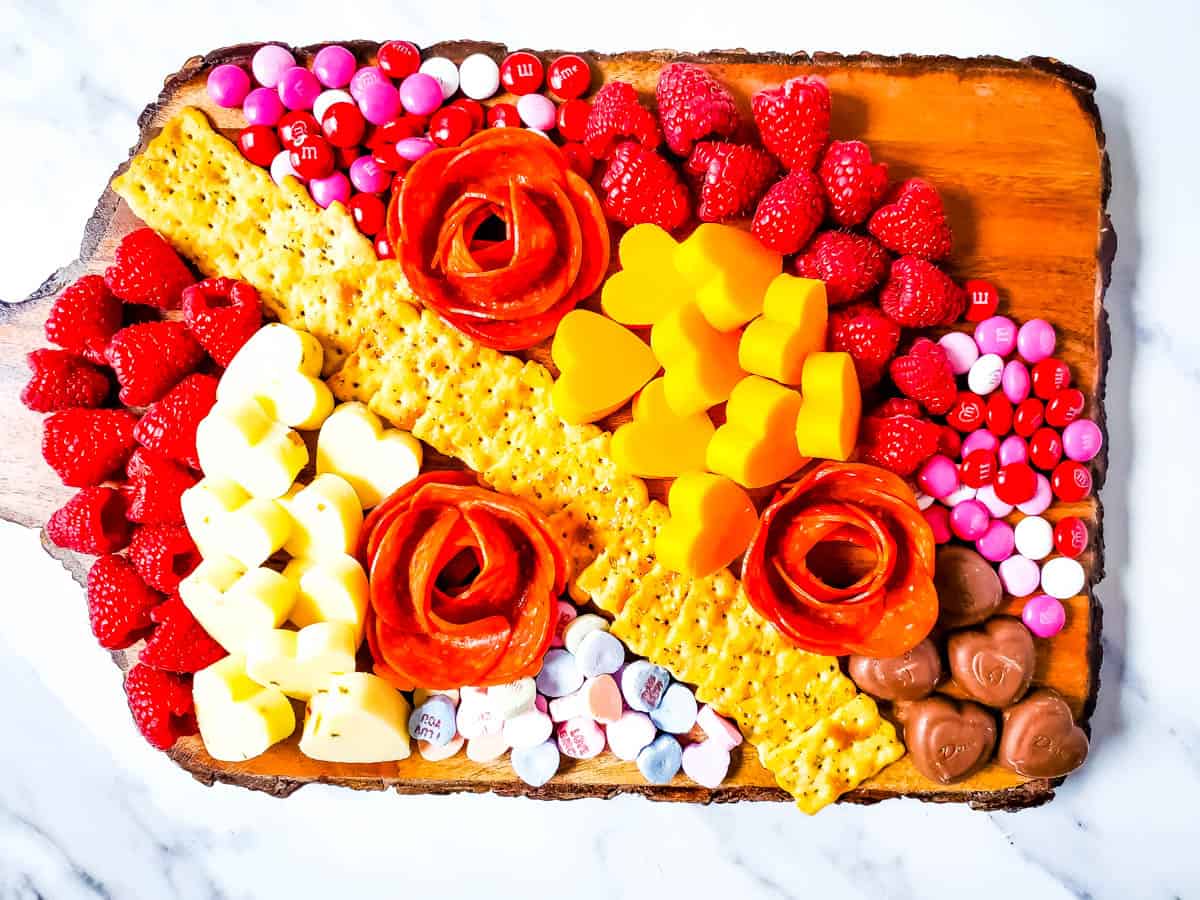 A wooden board with crackers, sliced cheese, pepperoni shaped as roses, raspberries, heart-shaped cheese, various chocolates, and red, pink, and white candy.