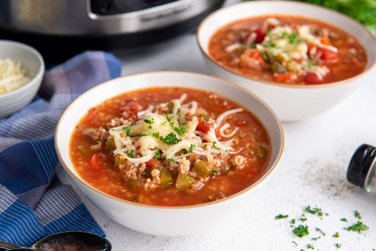 Two bowls of soup topped with shredded cheese and parsley, placed on a white surface with a blue napkin and a slow cooker in the background.
