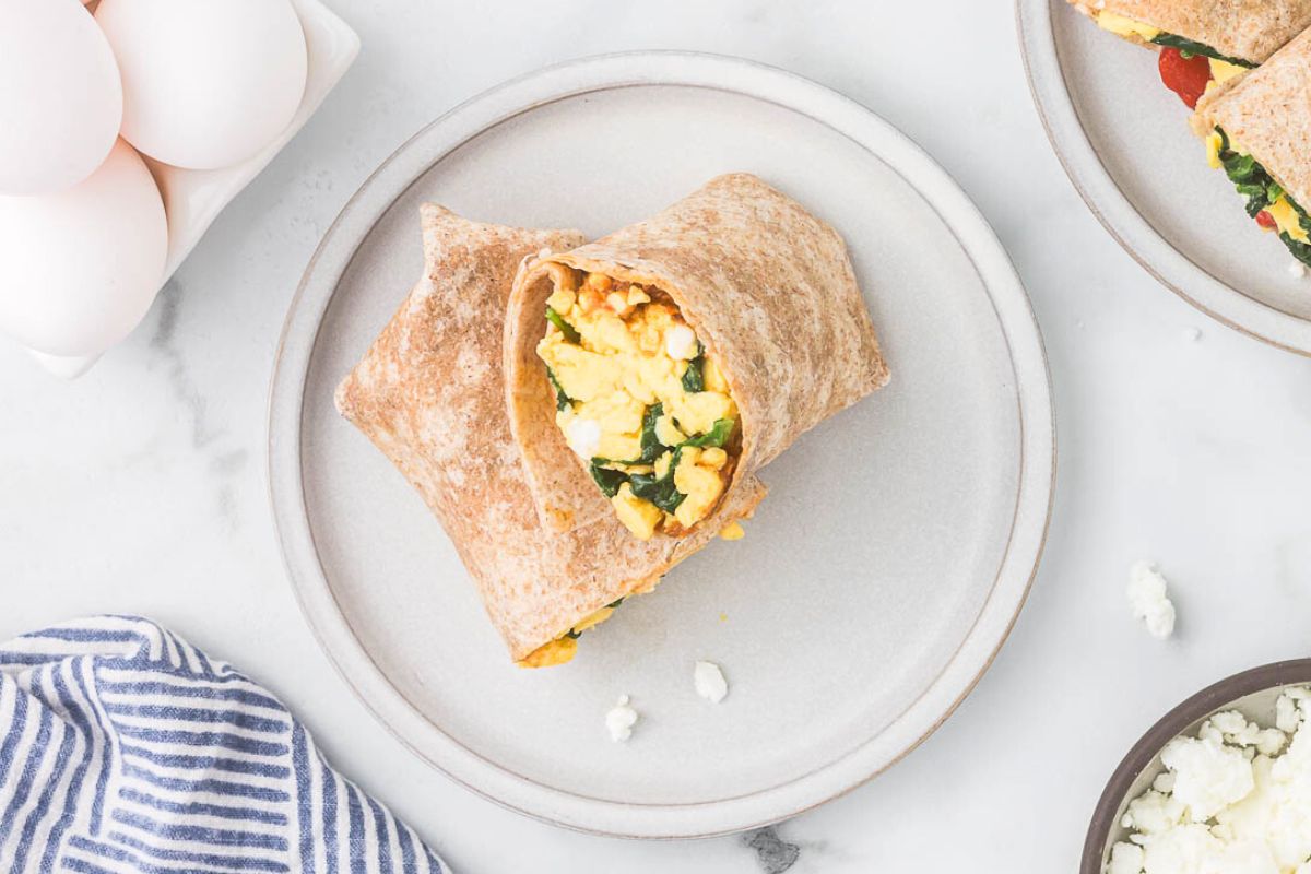 A whole wheat breakfast burrito filled with scrambled eggs and spinach sits on a white plate, with a bowl of eggs and a striped napkin nearby.
