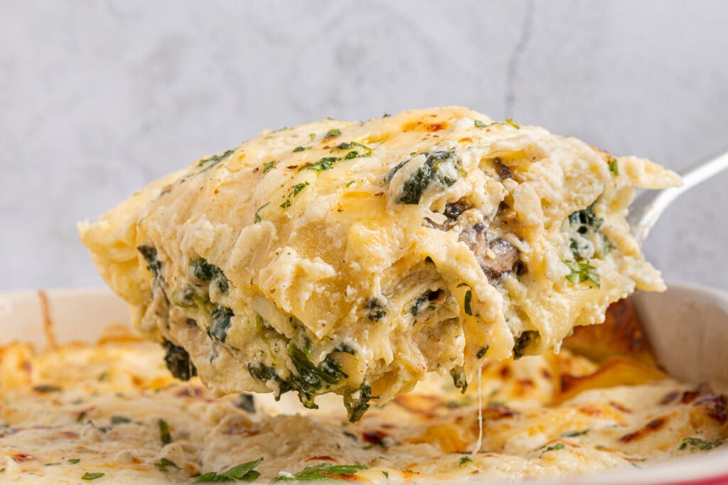 A close-up of a serving of creamy spinach and mushroom lasagna being lifted out of a baking dish with melted cheese on top.