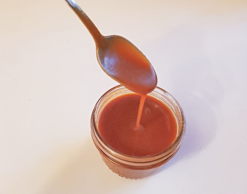 A spoon drizzling caramel sauce into a small glass jar filled with caramel sauce, set on a white surface.