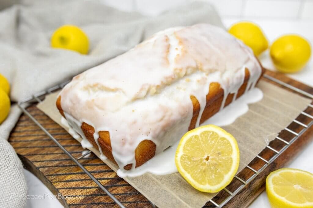 A loaf of lemon bread with white icing sits on parchment paper atop a cooling rack, surrounded by fresh lemons, one sliced in half.