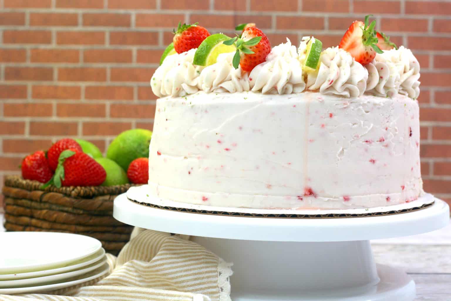 A frosted layer cake topped with whipped icing, strawberries, and lime slices sits on a white cake stand. A basket of limes and strawberries is in the background.