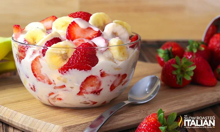 A glass bowl filled with sliced strawberries and bananas mixed in creamy yogurt sits on a wooden board with a spoon and fresh strawberries beside it.