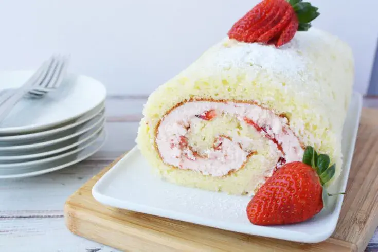 A sliced strawberry Swiss roll cake with cream filling, garnished with fresh strawberries, sits on a white plate next to a stack of plates and forks.