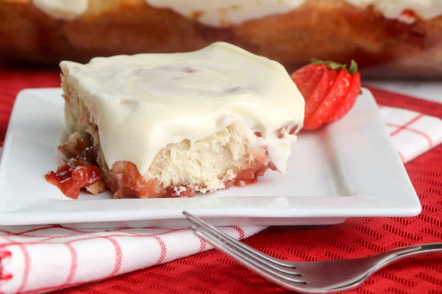 A square piece of frosted strawberry pastry on a white plate, garnished with a sliced strawberry, with a fork and red checkered napkin nearby.