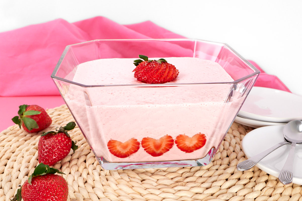 A clear glass bowl filled with pink strawberry mousse, topped and decorated with fresh strawberry slices, sits on a woven placemat next to whole strawberries, plates, and spoons.