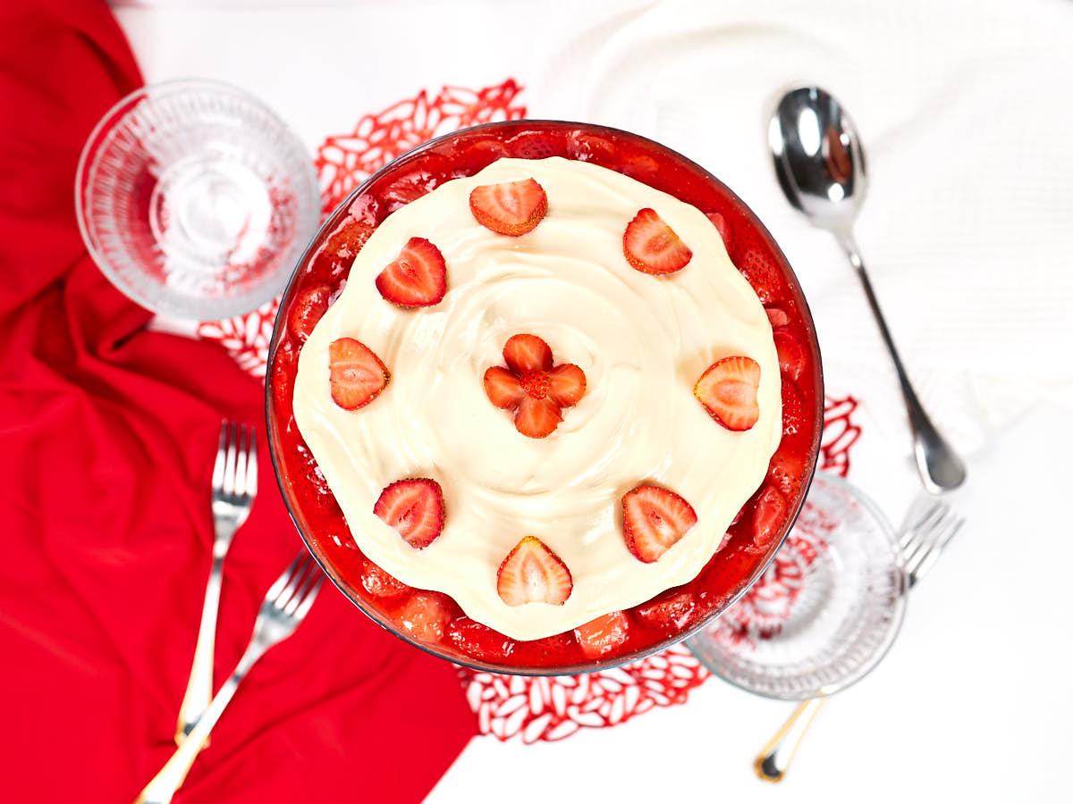 Overhead view of a trifle dessert topped with cream and sliced strawberries, surrounded by glass bowls, forks, a spoon, and red and white linens.