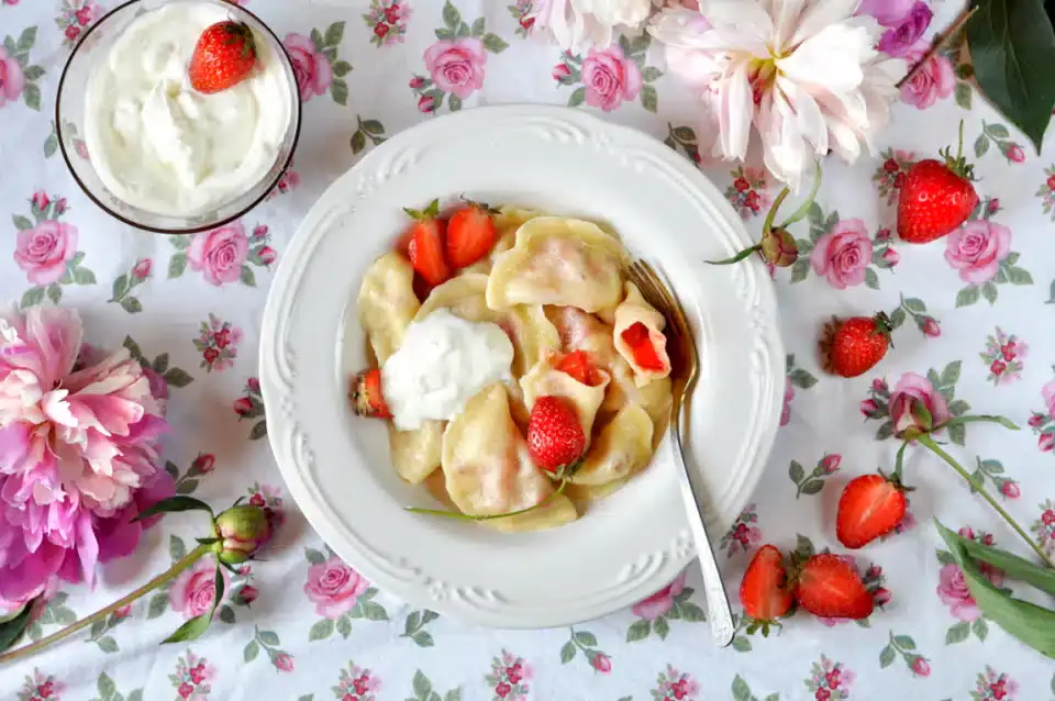 A plate of dumplings with strawberries and cream, garnished with fresh strawberries, sits on a floral tablecloth next to a bowl of cream and pink flowers.