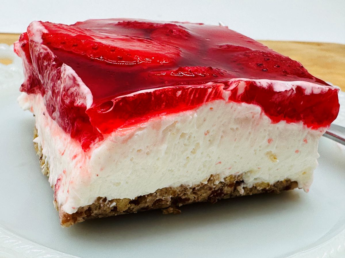 A slice of dessert with a crumbly base, thick creamy layer, and a gelatinous strawberry topping on a white plate.