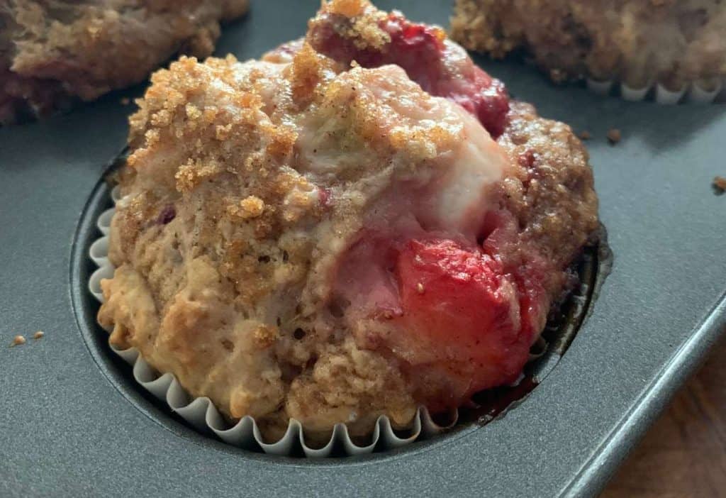A close-up of a baked strawberry muffin in a white paper liner, sitting in a dark muffin tin.