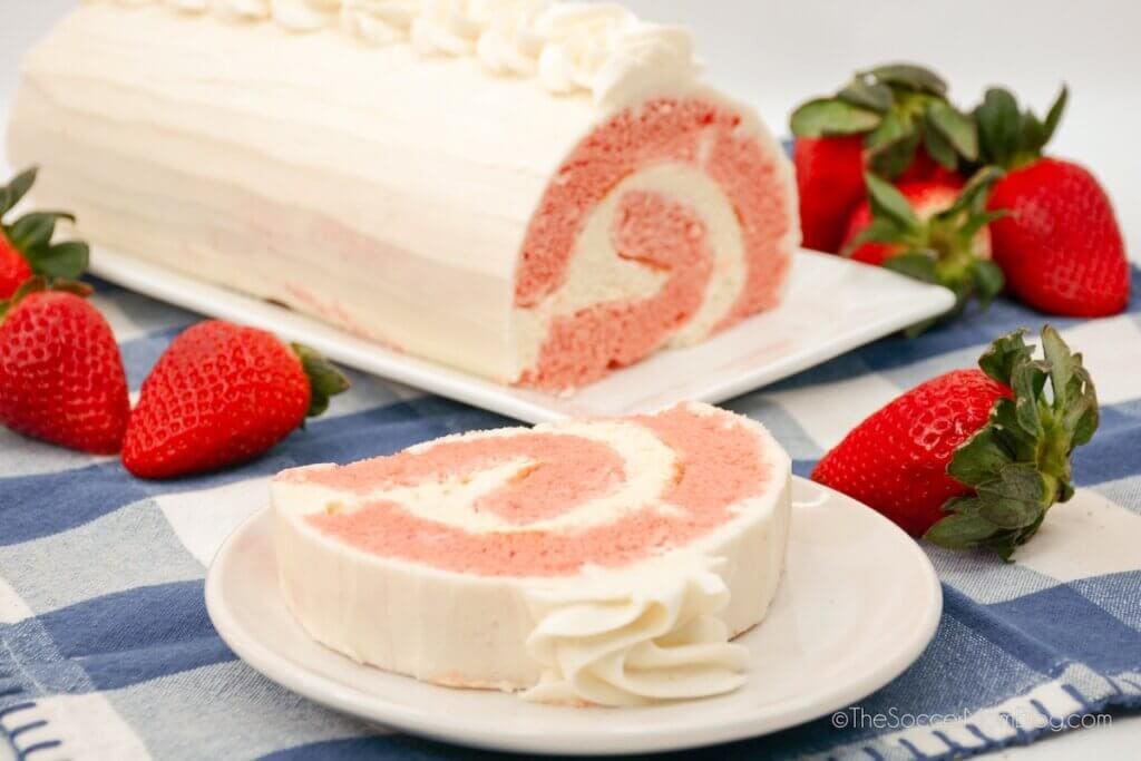 A slice of strawberry roll cake with white frosting on a plate, whole cake and fresh strawberries in the background, set on a blue and white checkered cloth.