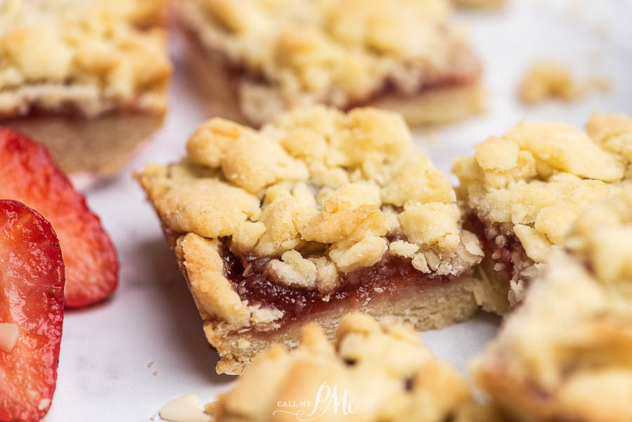Close-up of crumbly strawberry jam bars on a white surface, with fresh strawberry slices visible beside them.