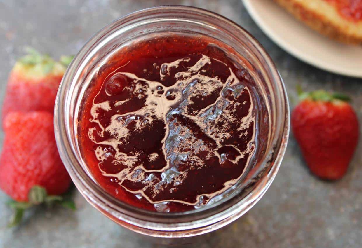 A close-up view of a glass jar filled with strawberry jam, with two whole strawberries and part of a slice of bread visible in the background.