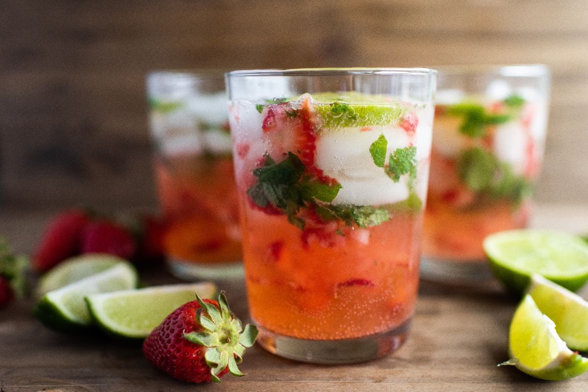 Three glasses of a strawberry and lime drink with ice and mint sit on a wooden surface, surrounded by fresh strawberries and lime wedges.