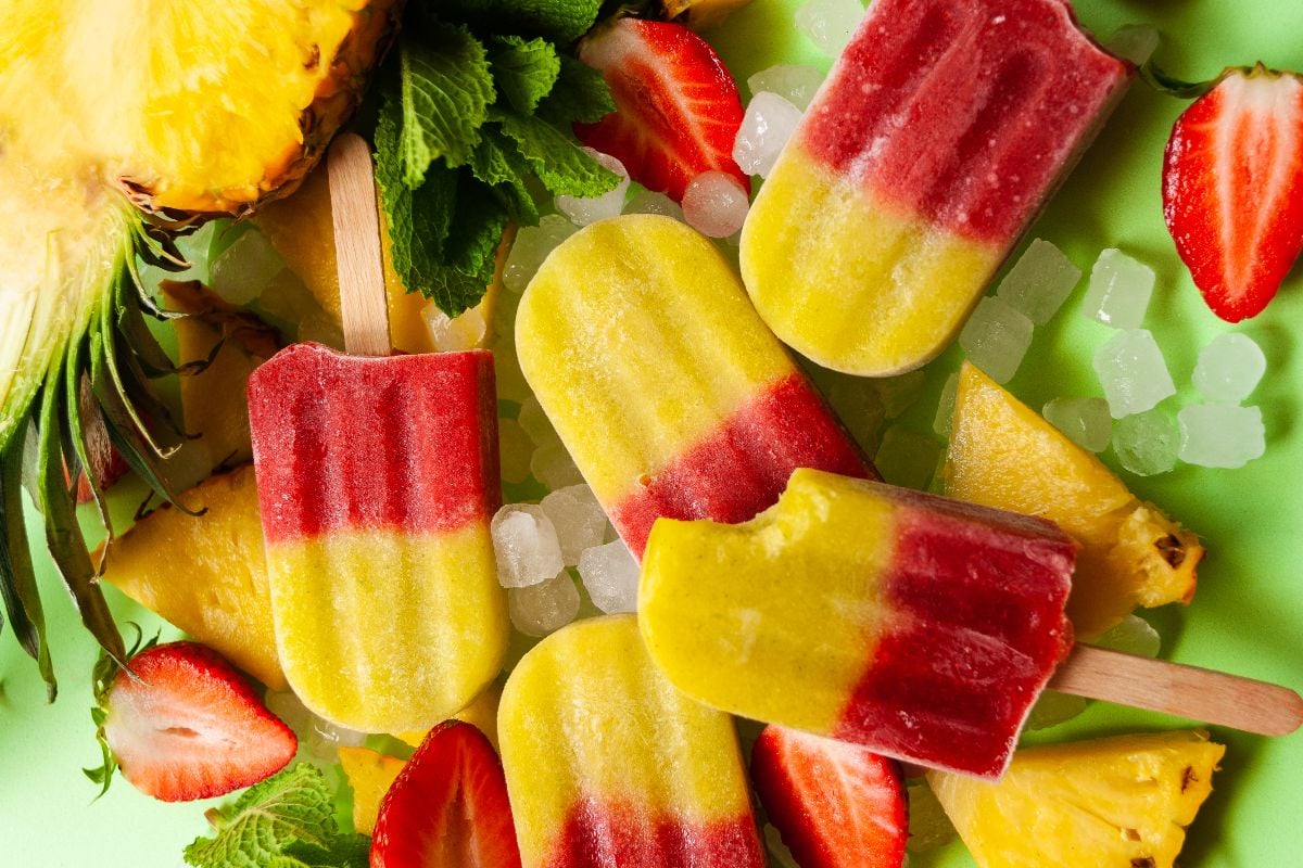 Assorted red and yellow popsicles surrounded by pineapple slices, strawberries, mint leaves, and ice cubes on a light green background. One popsicle has a bite taken out.
