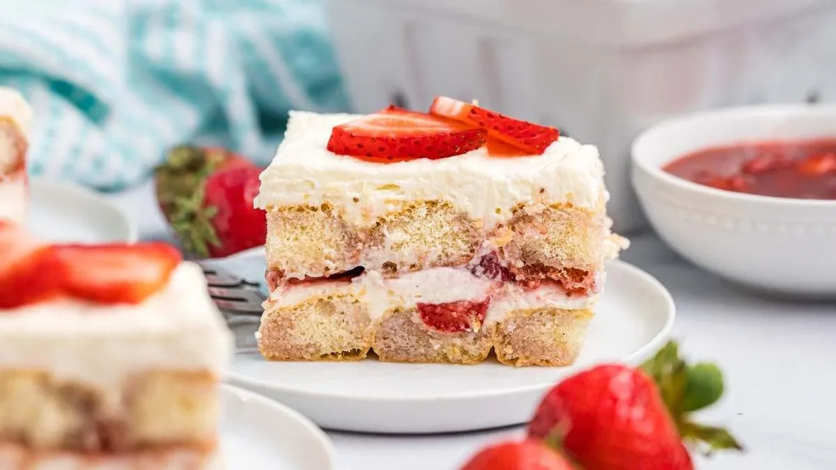 A slice of layered strawberry shortcake with cream topping and sliced strawberries on a white plate, with whole strawberries and a bowl of sauce in the background.