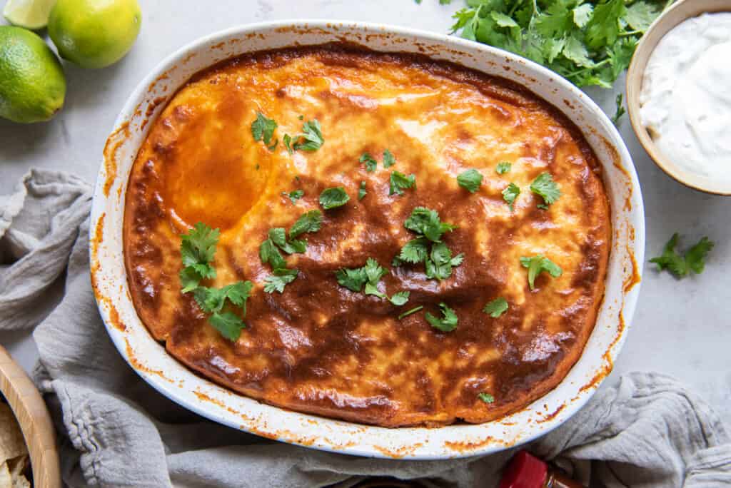 A baked casserole topped with melted cheese and garnished with fresh cilantro, with limes, cilantro, and sauce visible nearby.