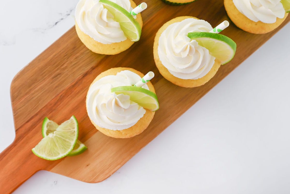 Four cupcakes with white frosting and lime wedges on top, arranged on a wooden board with additional lime slices beside them.