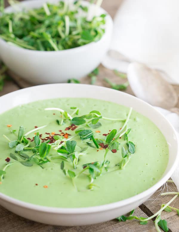 A bowl of creamy green soup garnished with fresh greens and red pepper flakes, with a spoon and a bowl of greens in the background.